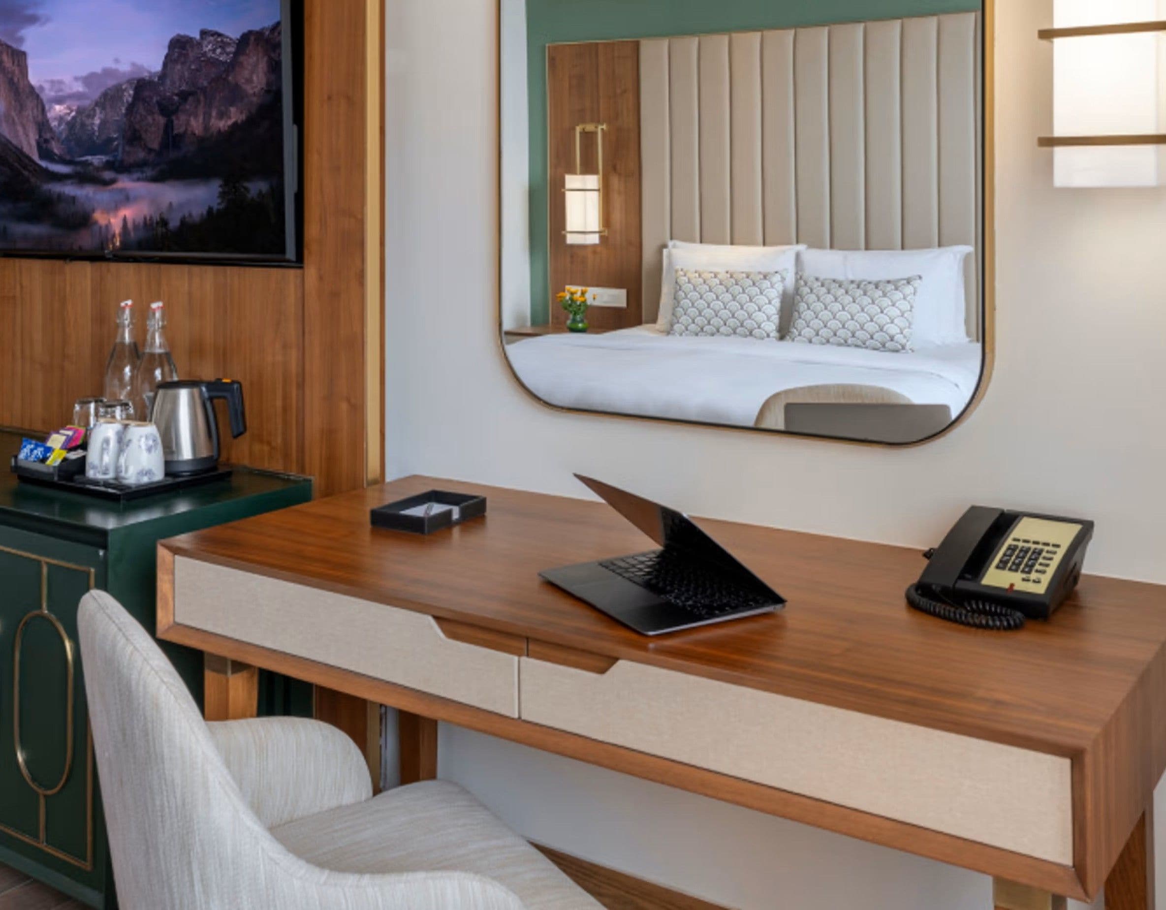 Habitación de hotel con un escritorio de madera, una computadora portátil, un teléfono y una cama con espejo en la cabecera.