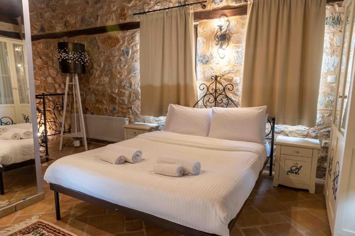 Un dormitorio con pared de piedra, una cama con ropa de cama blanca, una mesita de noche, una lámpara y una ventana con cortinas.