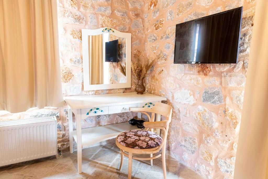 Ein Zimmer mit Steinwand, weißem Schreibtisch, einem Sessel mit Blumenkissen, einem Spiegel und einem an der Wand befestigten Flachbildfernseher.