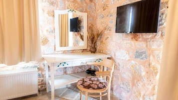 Ein Zimmer mit Steinwand, weißem Schreibtisch, einem Sessel mit Blumenkissen, einem Spiegel und einem an der Wand befestigten Flachbildfernseher.