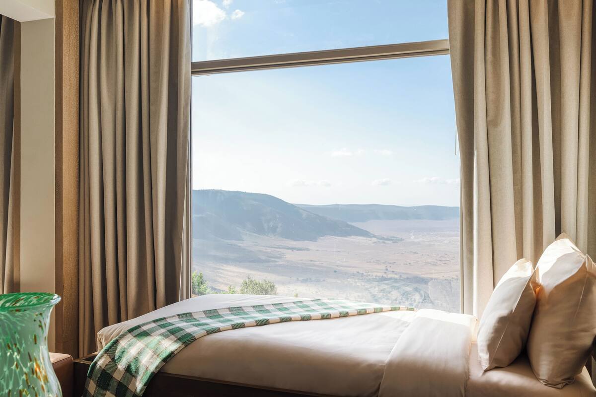 A hotel room with a large window, a bed with a checkered blanket, and a view of a mountainous landscape.