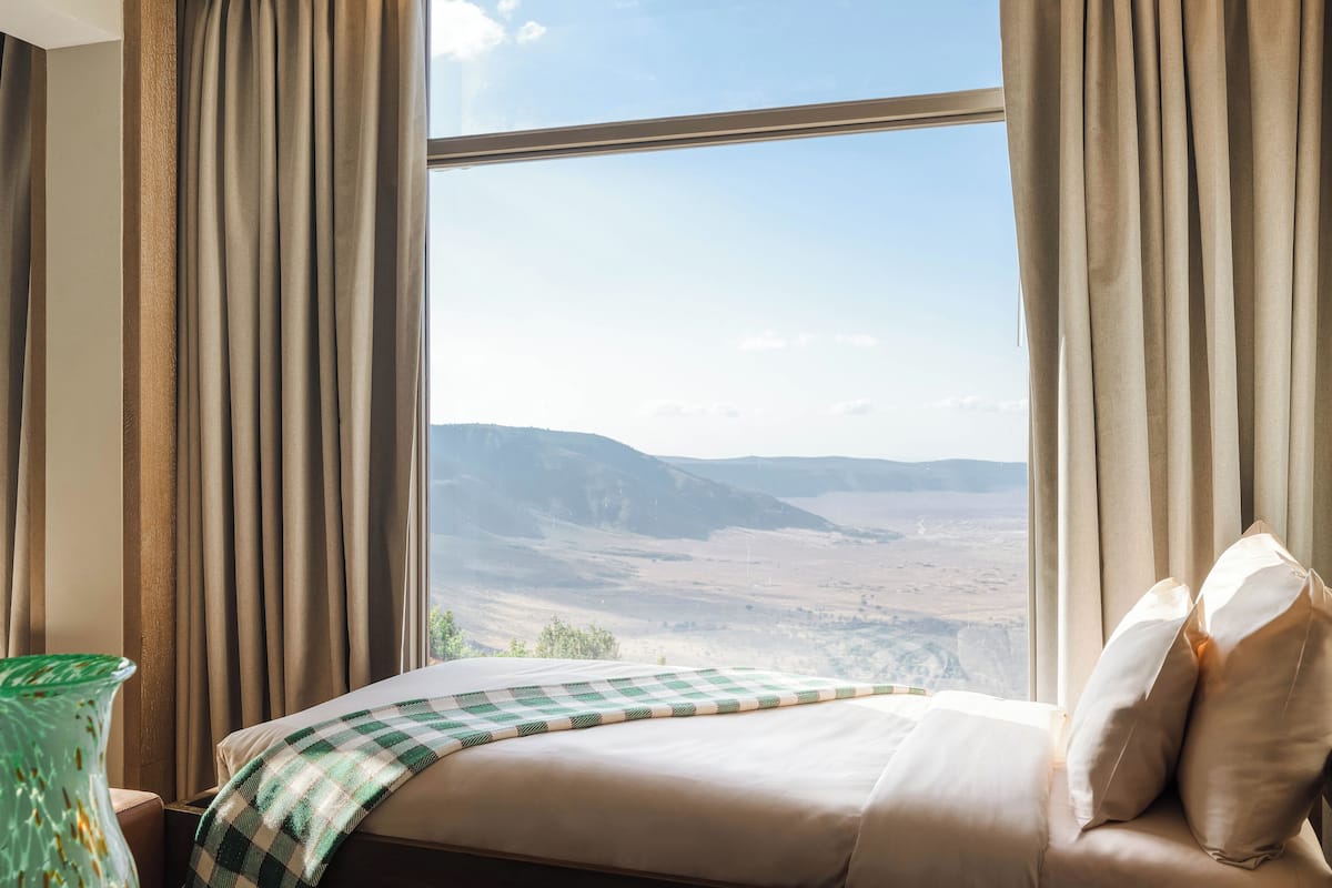 A hotel room with a large window, a bed with a checkered blanket, and a view of a mountainous landscape.