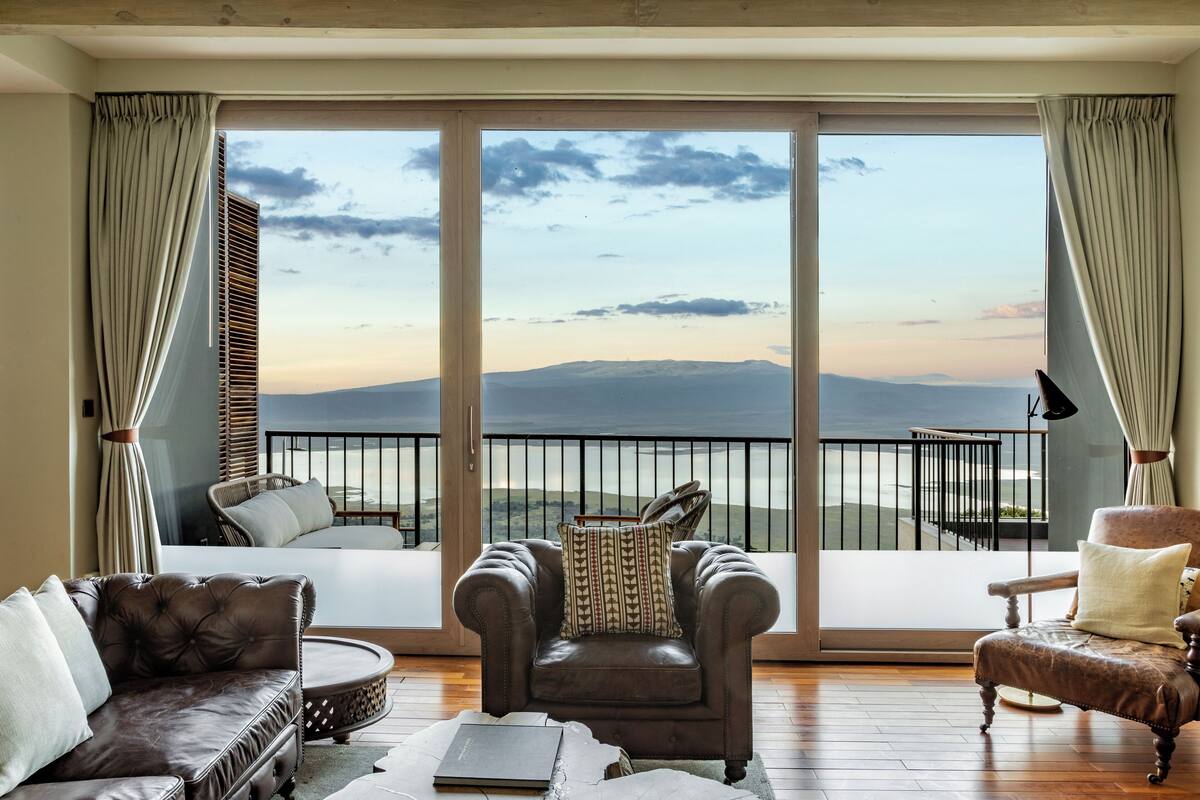 A room with a large sliding glass door leading to a balcony with a view of mountains. There are two leather armchairs and a sofa with cushions.