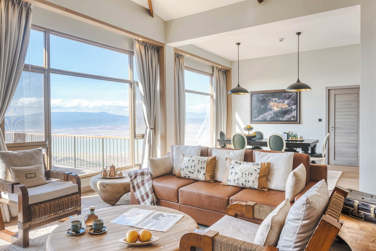 A spacious living room with a large window offering a view of the mountains, a cozy brown leather sofa, and a dining area with a table and chairs.