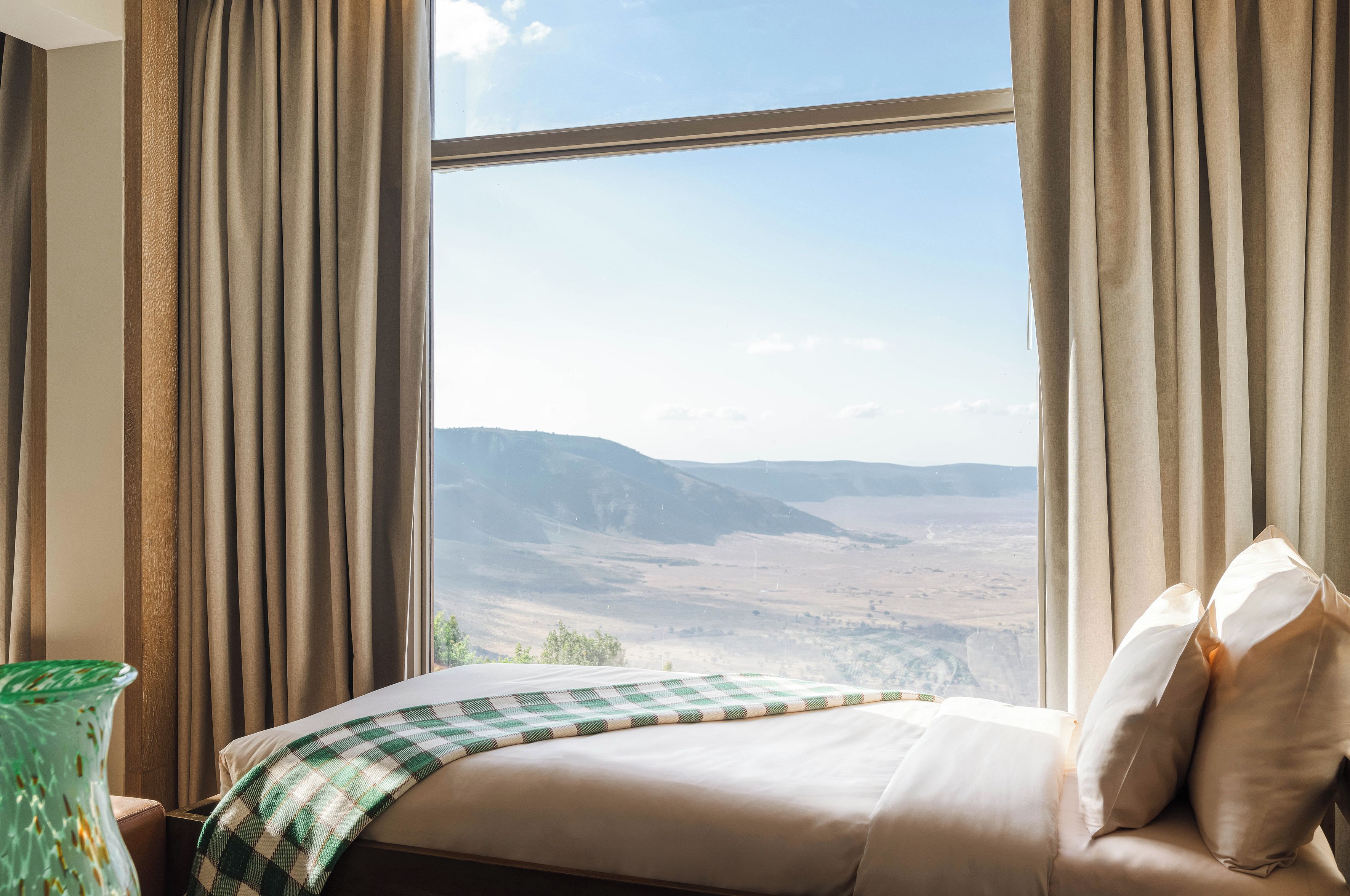 A hotel room with a large window, a bed with a checkered blanket, and a view of a mountainous landscape.