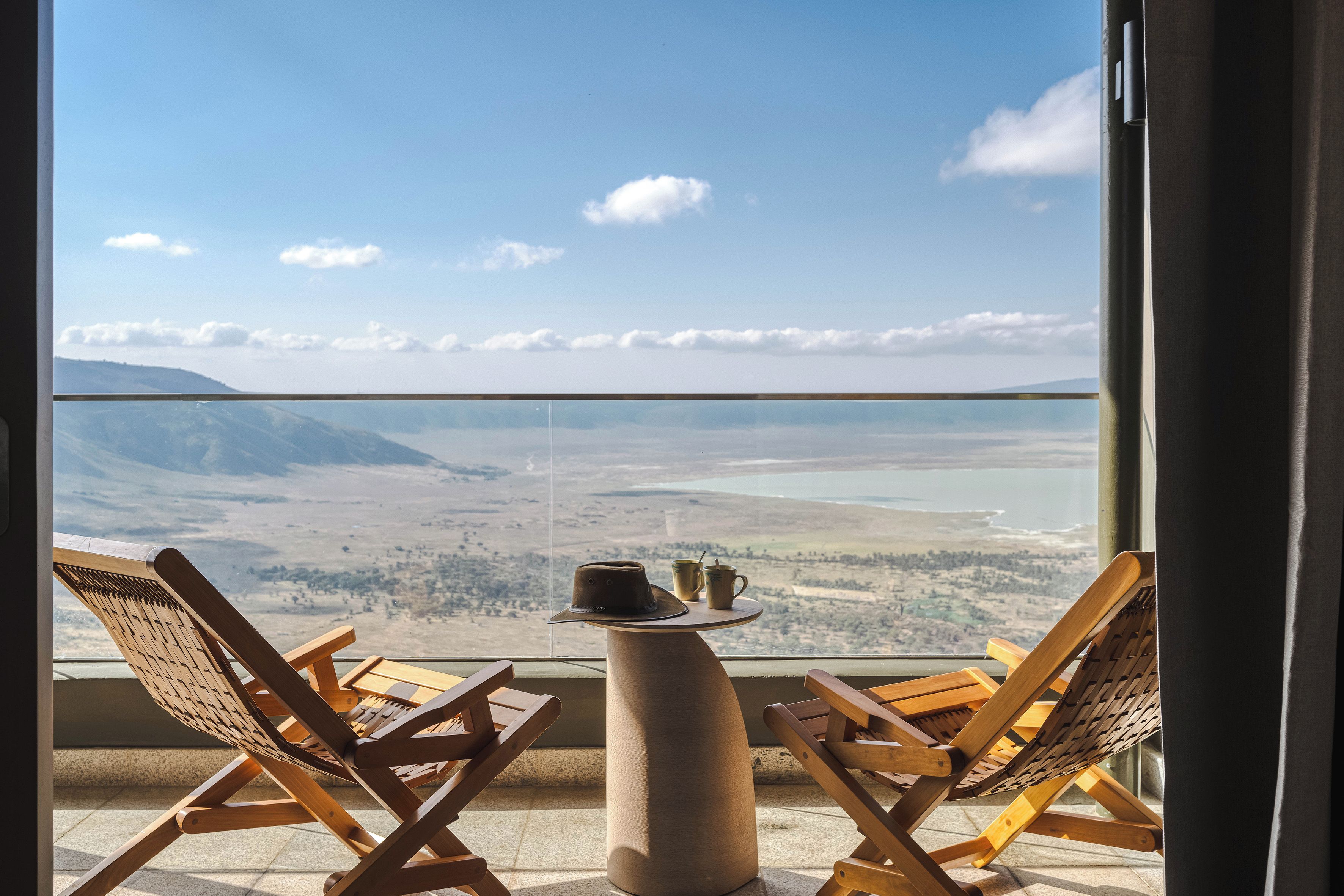 A balcony with two wooden lounge chairs and a small table, overlooking a scenic landscape with mountains and a body of water.