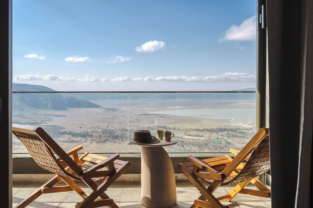 A balcony with two wooden lounge chairs and a small table, overlooking a scenic landscape with mountains and a body of water.