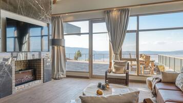 A spacious living room with a large TV, a fireplace, and a balcony with a view of the sea.