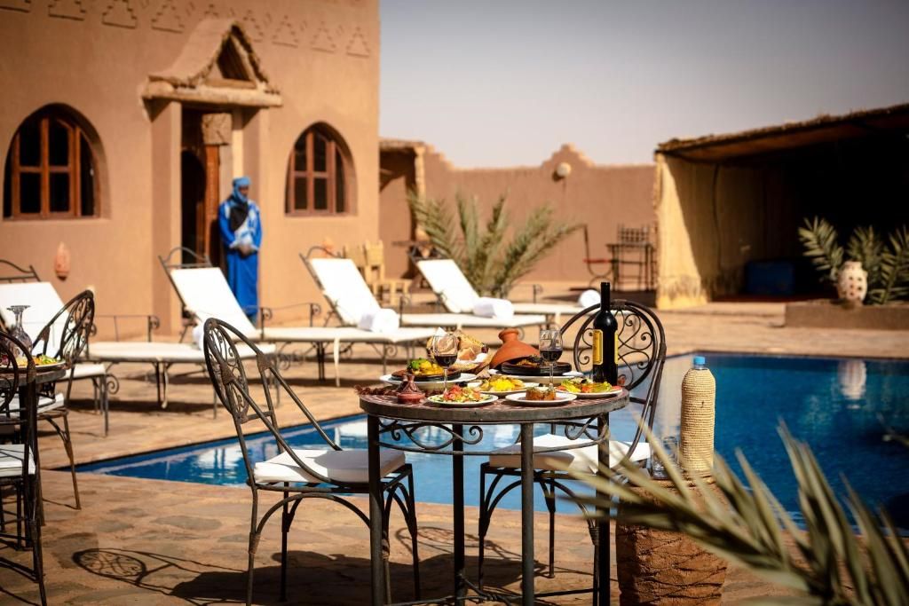 Un área de comedor junto a la piscina con sillones reclinables, una mesa dispuesta con comida y bebidas, y un edificio con ventanas arqueadas al fondo.