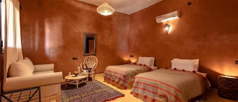 Quarto com duas camas, um sofá, uma mesa pequena, uma cadeira, luminária pendente e aparelho de ar condicionado.