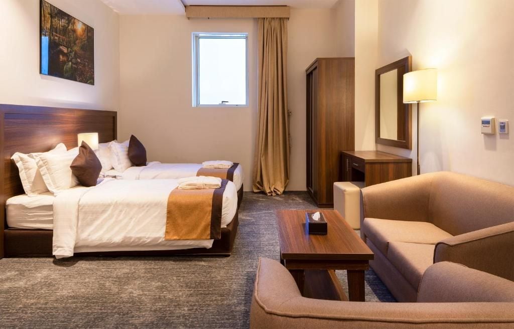 A hotel room with two beds, a sofa, a table, a lamp, and a window with curtains.