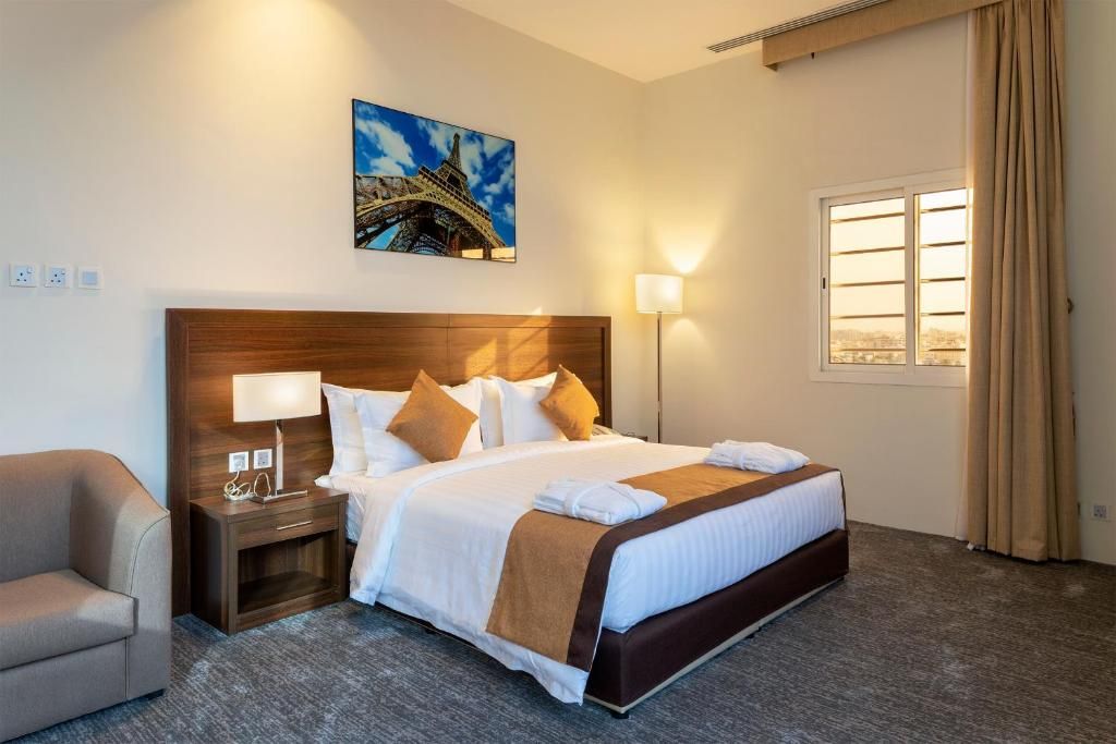 Habitación de hotel con una cama grande, una mesita de noche con lámpara, una silla, un ventanal con cortinas y un cuadro de la Torre Eiffel.