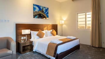 Habitación de hotel con una cama grande, una mesita de noche con lámpara, una silla, un ventanal con cortinas y un cuadro de la Torre Eiffel.
