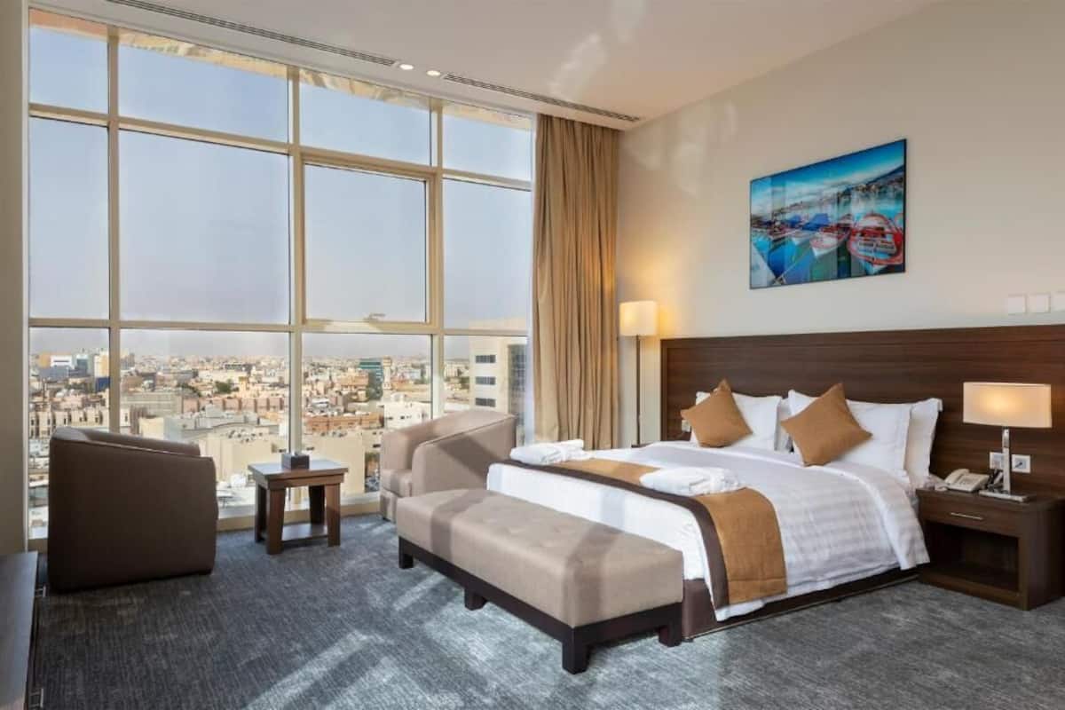 Habitación de hotel con un ventanal grande, una cama con ropa blanca, un cabecero marrón, una mesita de noche con lámpara y vistas a la ciudad.