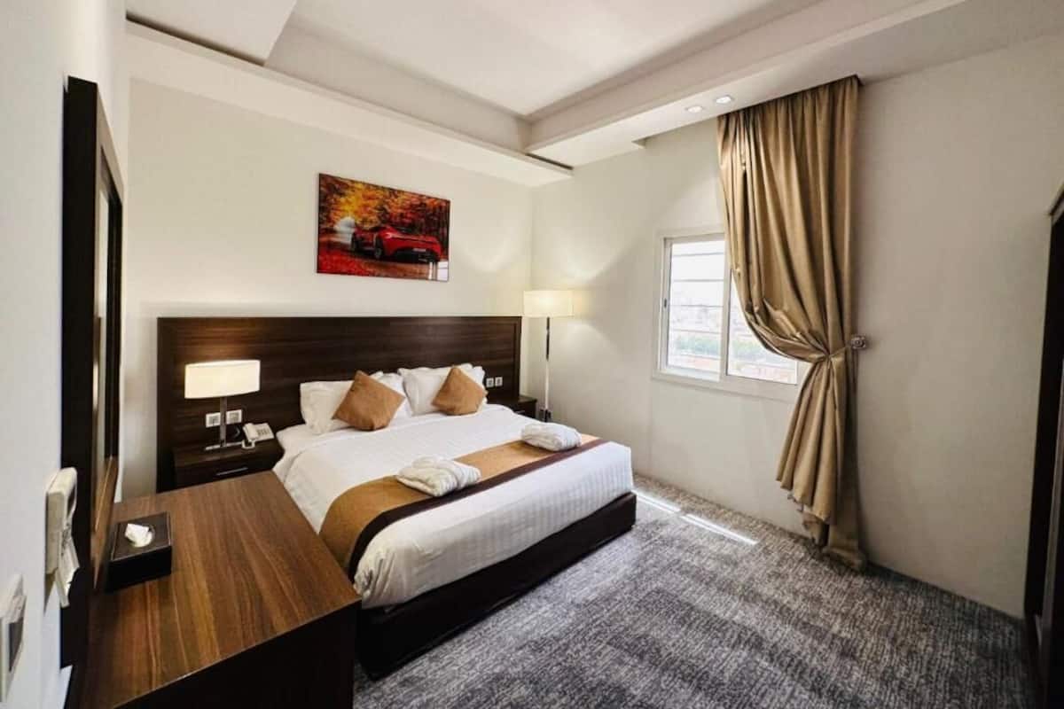 Una habitación de hotel con una cama grande, una mesita de noche de madera, un escritorio, una lámpara, un cuadro en la pared y una ventana con cortinas.