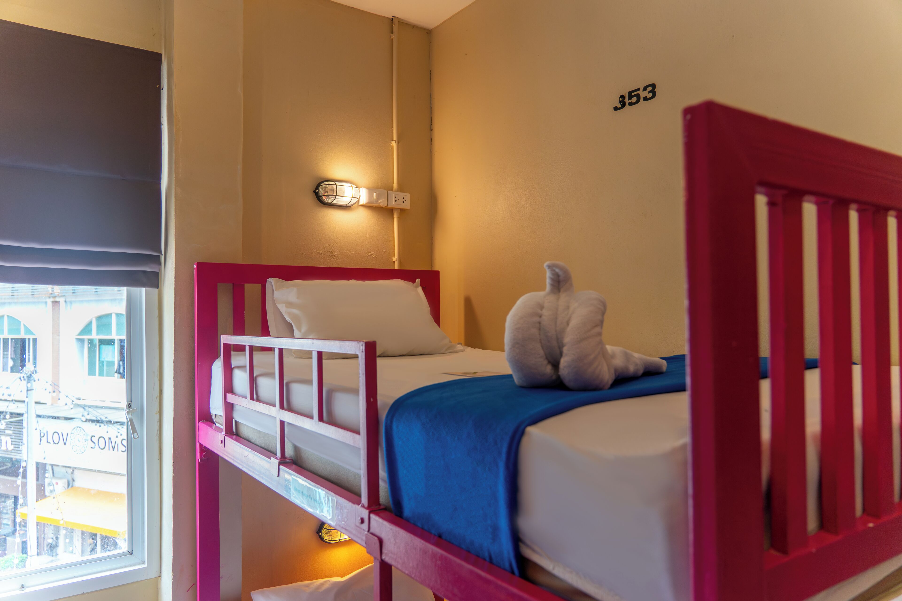 Una cama litera con cabecera roja, almohada blanca y manta azul. Una lámpara de pared está sobre la cama y se ve una ventana con persianas a la izquierda.