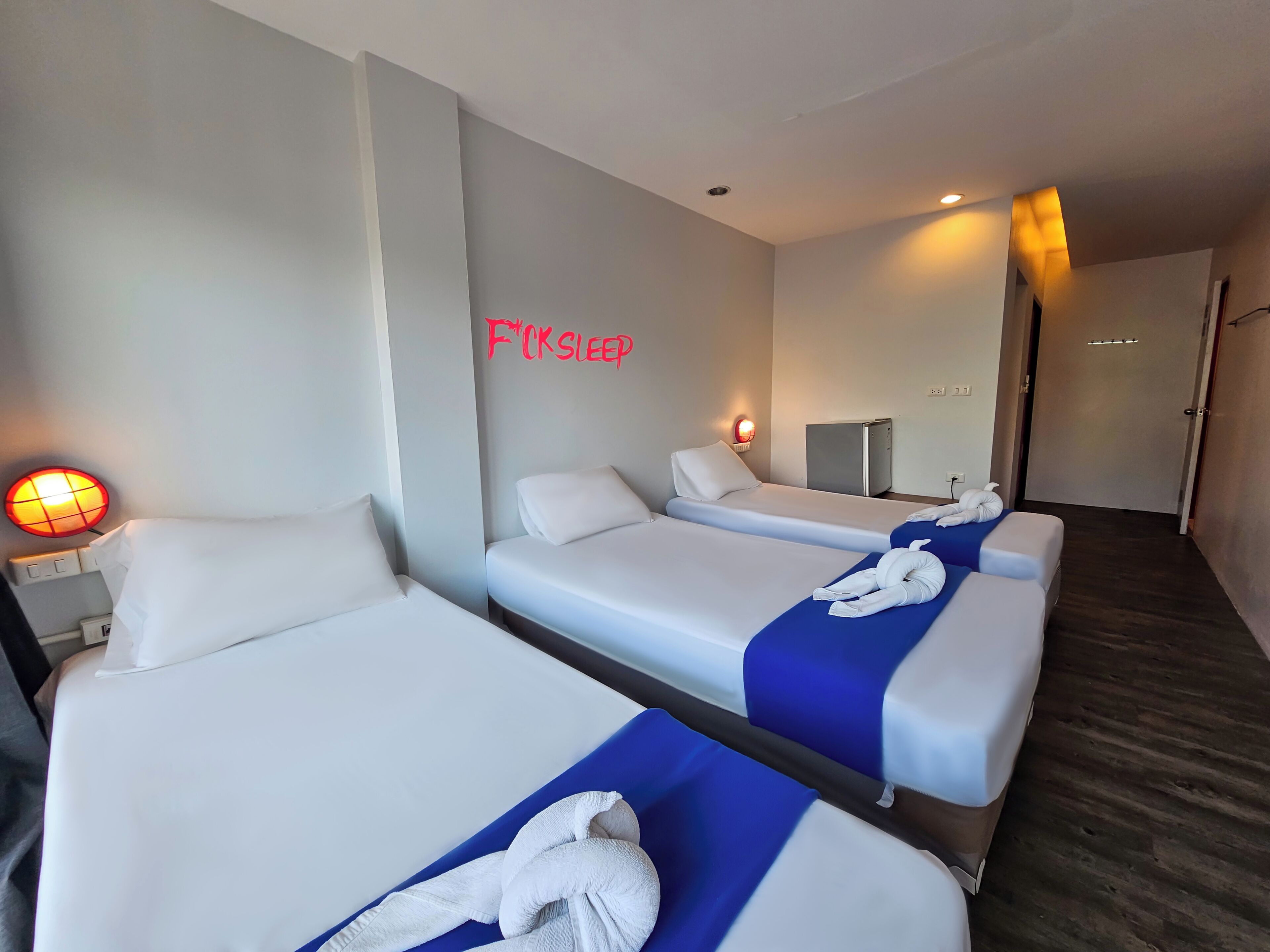 Una habitación de hotel con tres camas individuales, un escritorio con lámpara, un refrigerador pequeño y un espejo en la pared.