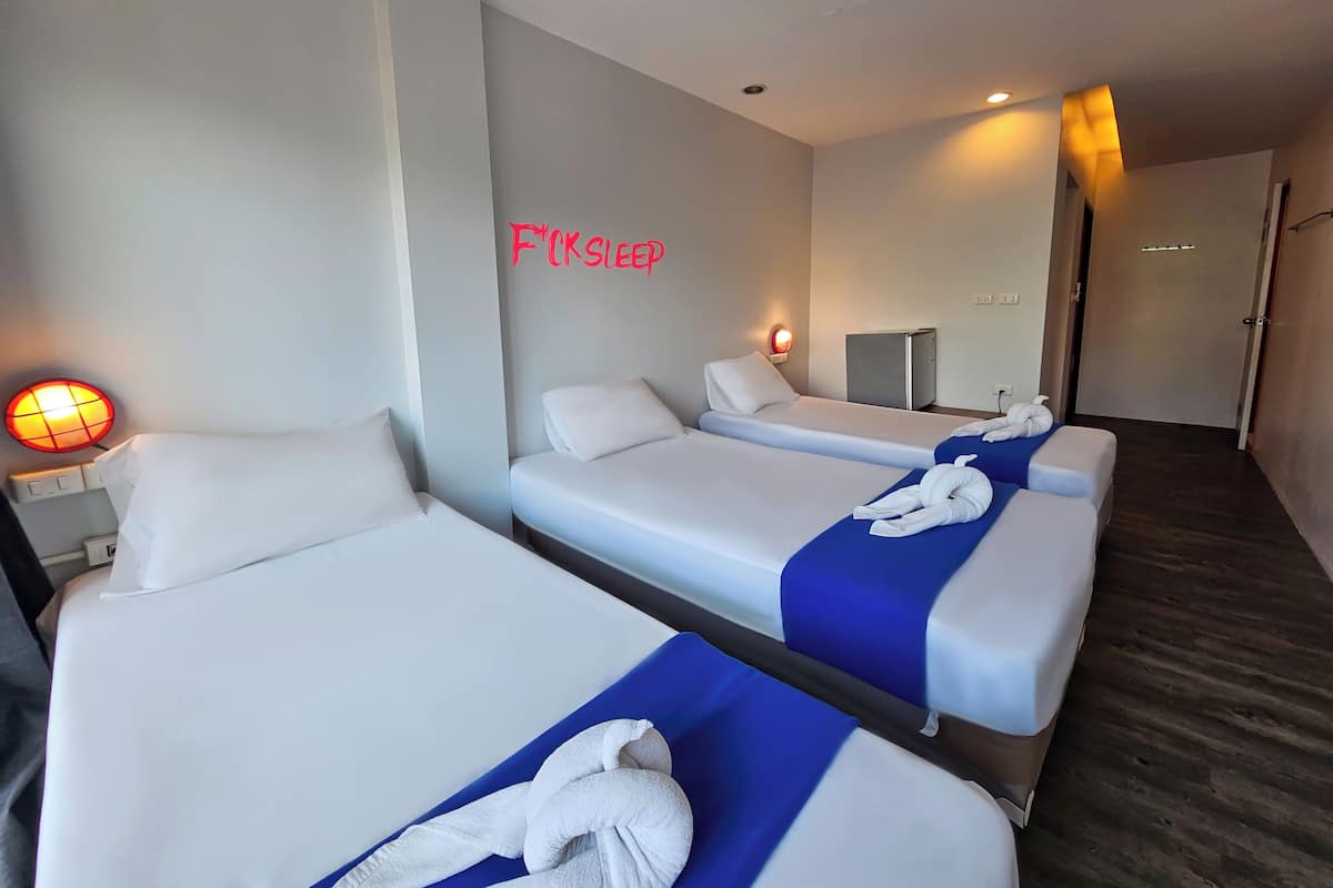Una habitación de hotel con tres camas individuales, un escritorio con lámpara, un refrigerador pequeño y un espejo en la pared.