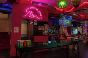 A neon sign reading 'If it swells, ride it,' a counter with a menu board, and a table with colorful cups. - Slumber Party Surf Kata (Karon)