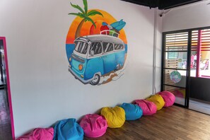 Une pièce dont un mur est décoré d’une fresque représentant un van bleu, une planche de surf, un palmier et un coucher de soleil. Des poufs colorés sont disposés le long du mur.