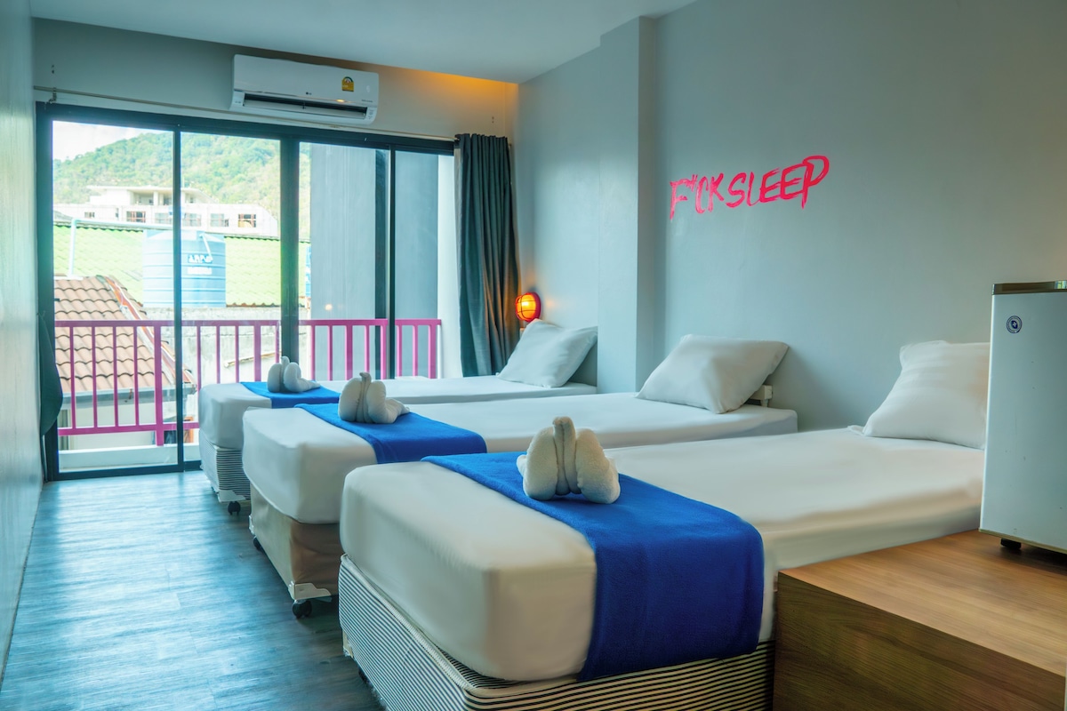 A hotel room with two beds, a balcony with a view of a building and greenery, and a wall with the text "FOKSLEEP".
