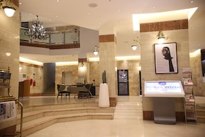 Vestíbulo de hotel con suelo de mármol, un mostrador de recepción, una escalera, una araña y un quiosco digital de información.