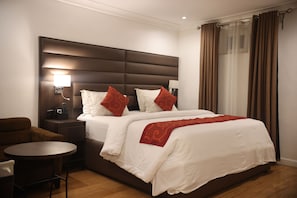 Habitación de hotel con una cama grande, una mesita de noche con lámpara, una pequeña mesa redonda y un sillón marrón.
