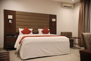 Una habitación de hotel con una cama grande, dos mesitas de noche, una lámpara, una silla, una pequeña mesa y un equipo de aire acondicionado.