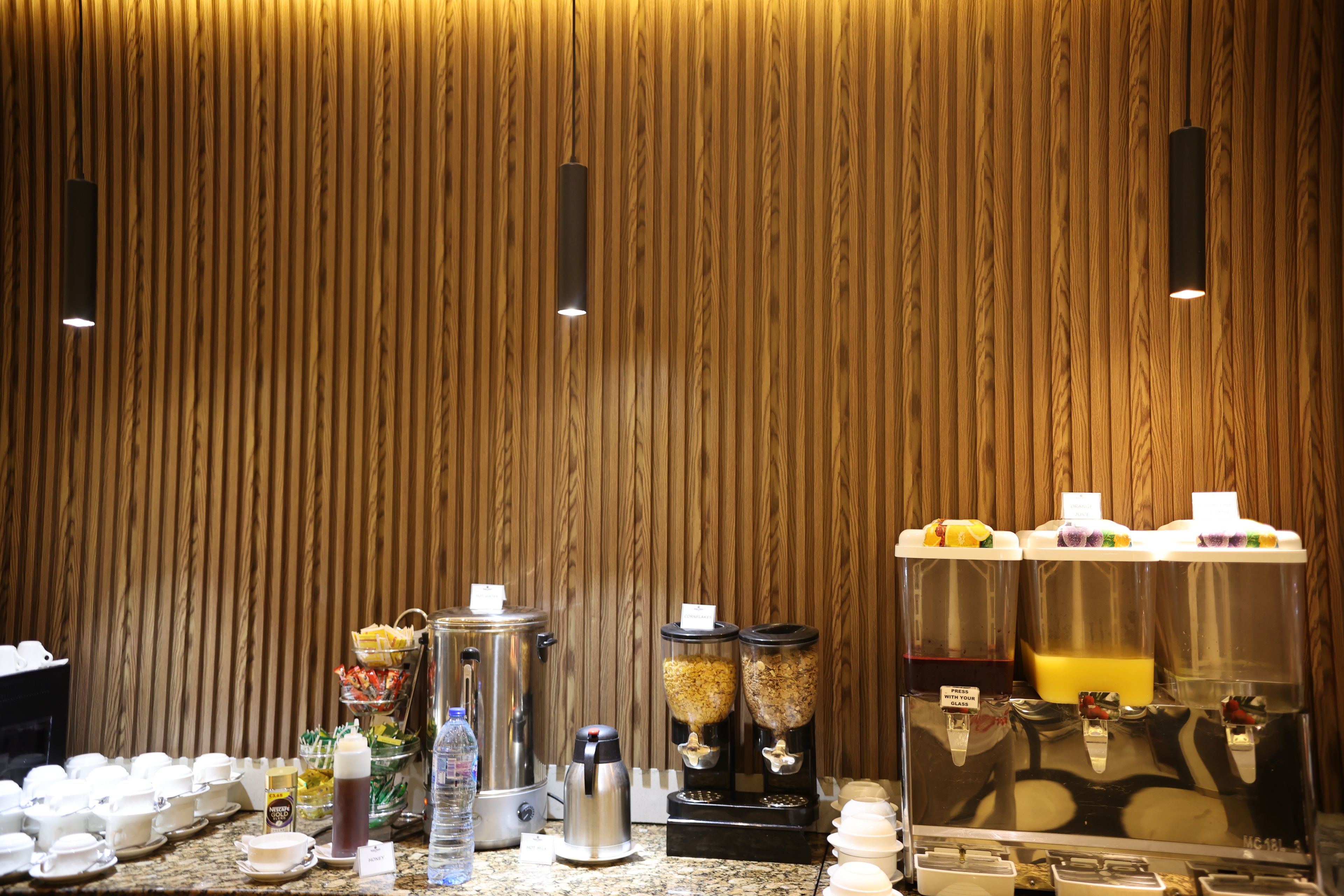 A breakfast bar with various food and drink dispensers, a coffee maker, and a water dispenser against a wooden wall.