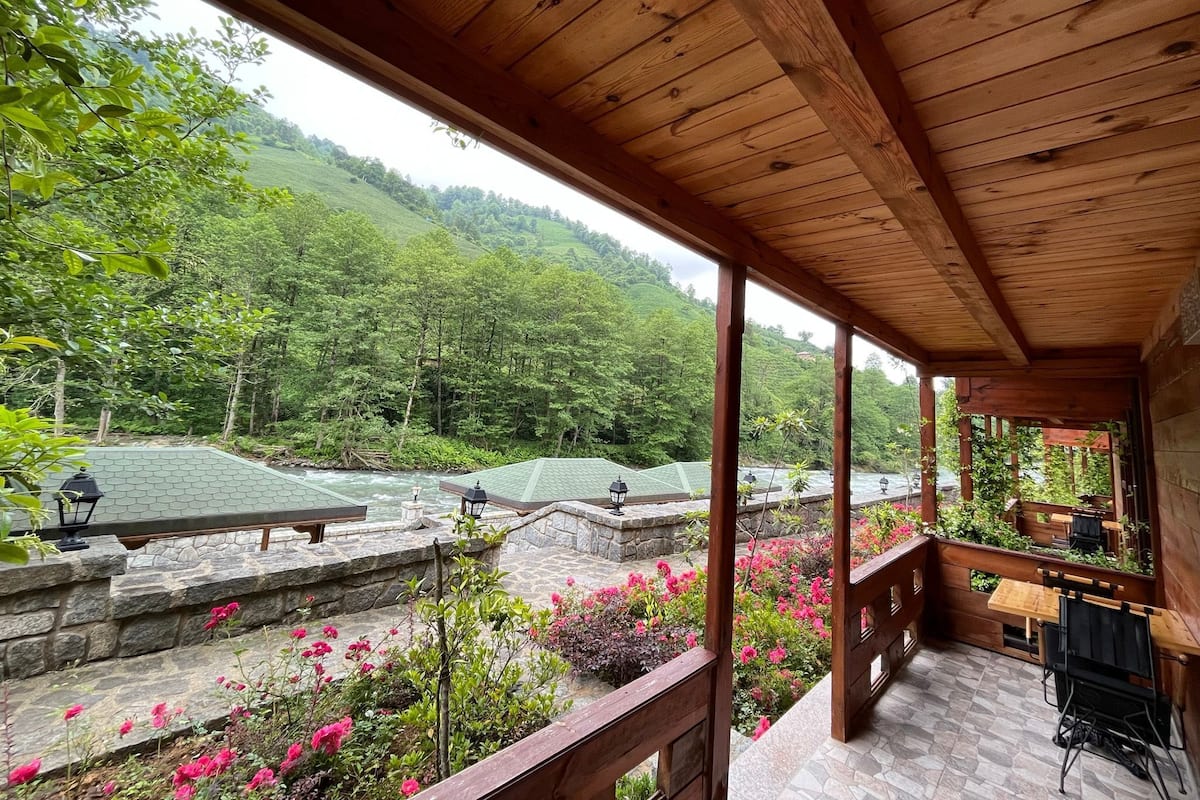 Eine Holzterrasse mit Steinzaun, die auf einen Garten mit rosa Blumen und Blick auf einen Fluss und bewaldete Hügel blickt.