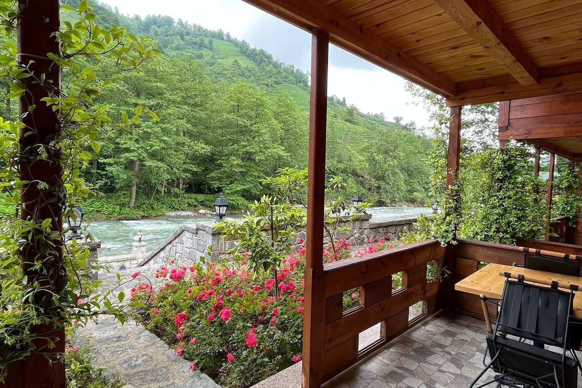 Eine überdachte Terrasse mit Steinböden, Holzmöbeln und Blick auf einen Fluss und bewaldete Hügel.