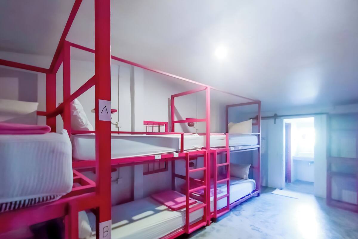 Beliches com estrutura rosa e roupa de cama branca, dispostos em linha, com uma porta à direita.