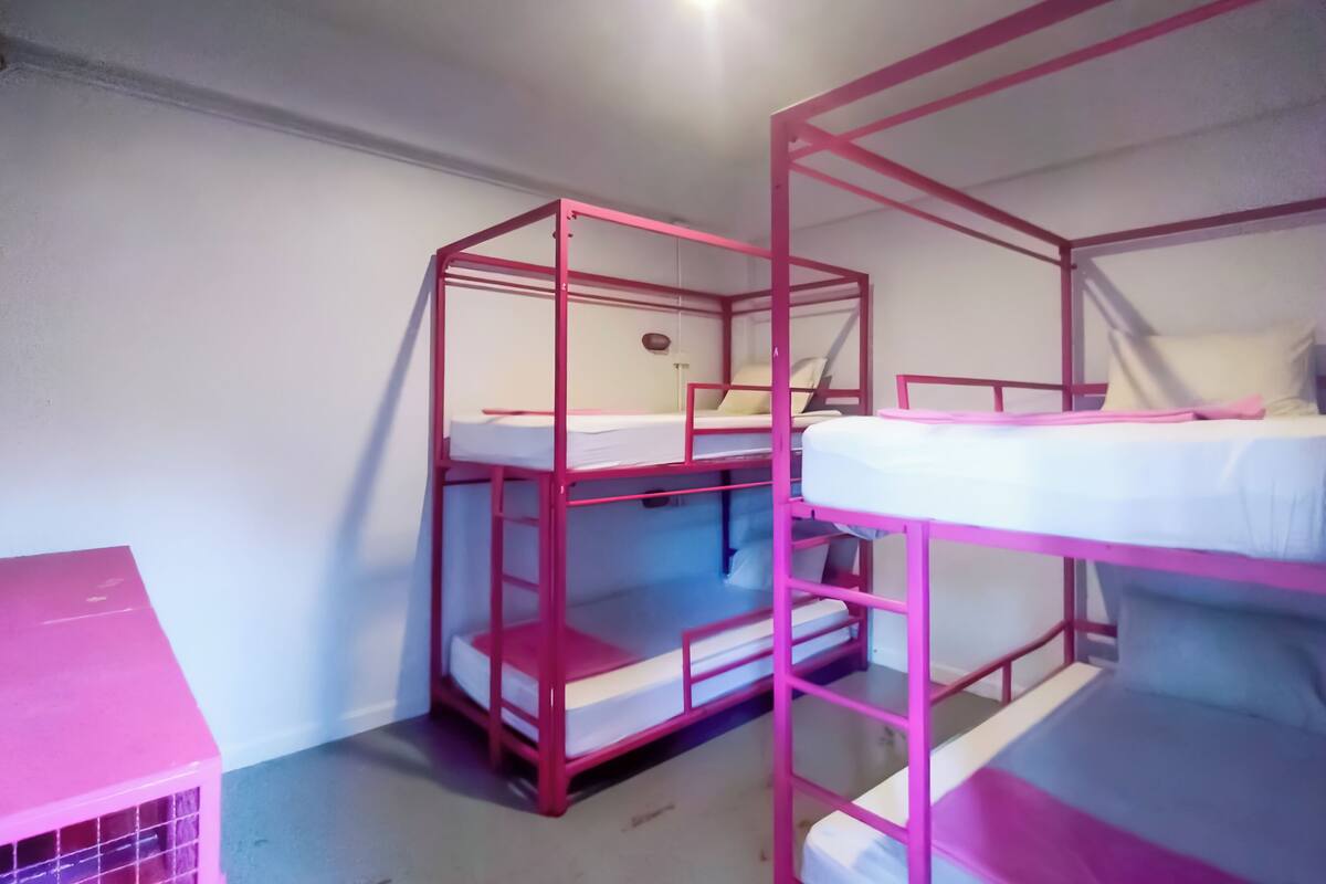 Quarto com beliche de estrutura metálica rosa, uma mesinha e prateleira fixada na parede.
