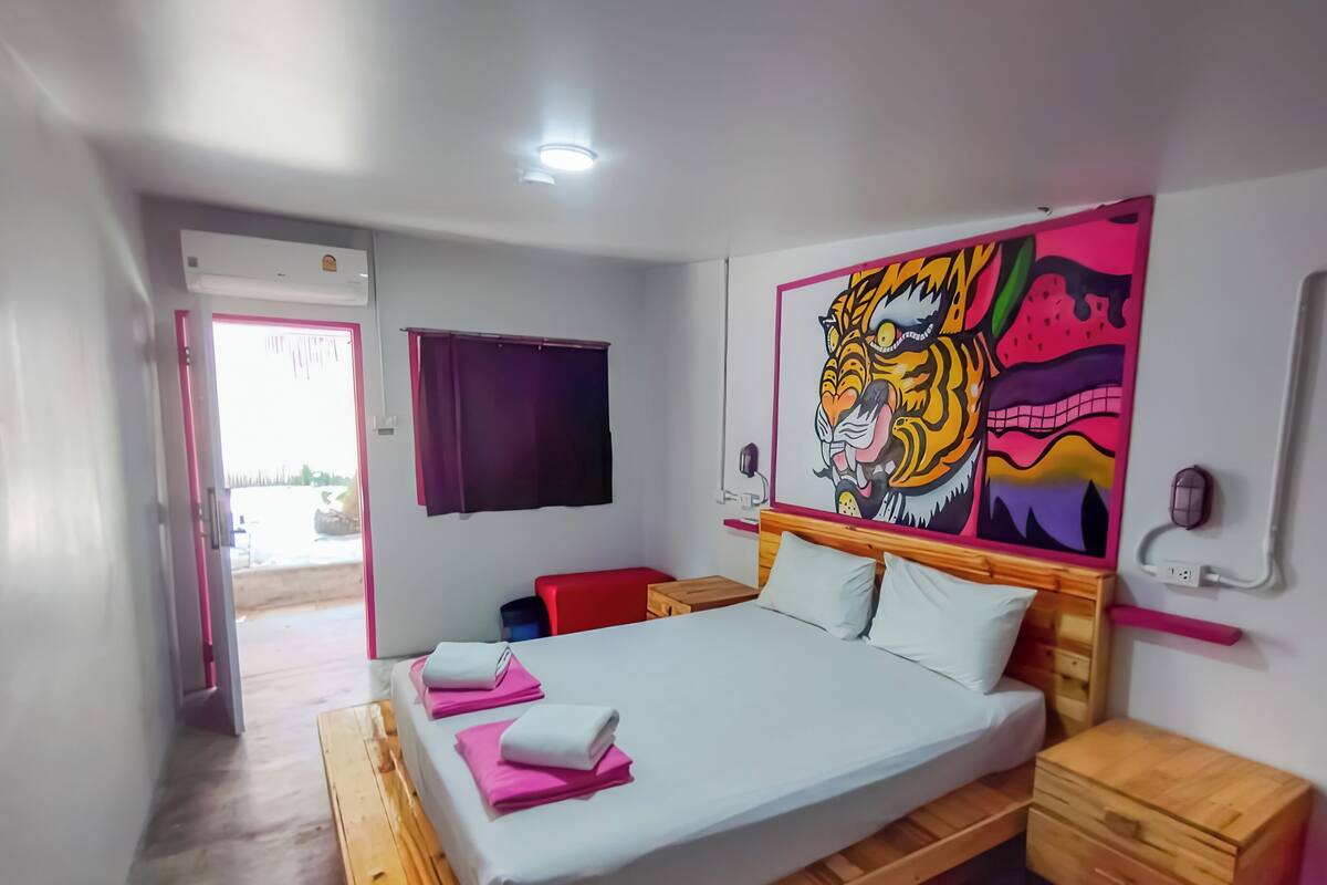 Um quarto com uma cama, mesa de cabeceira, cômoda de madeira, janela com cortinas e um mural na parede.