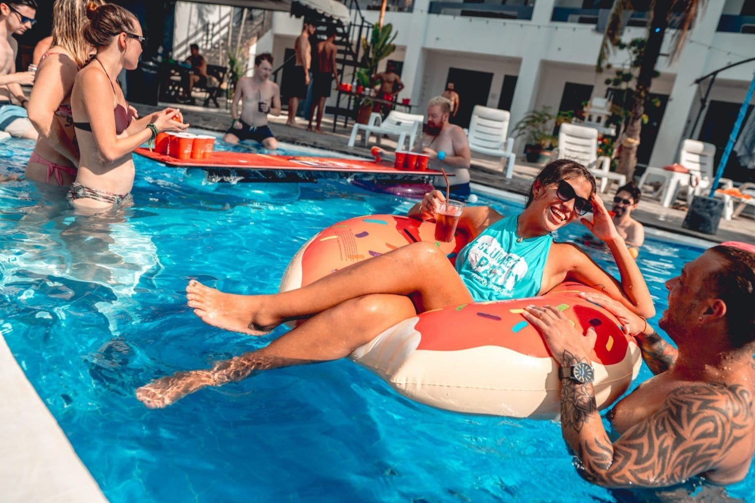 Zona de piscina con gente relajándose en flotadores, un área de sillones de descanso y un edificio de diseño moderno.