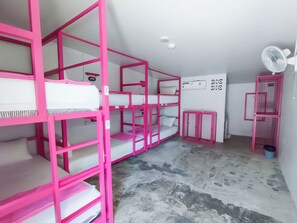 Camas litera con estructura rosa, un equipo de aire acondicionado blanco, un ventilador blanco y una pequeña mesa con un bote de basura azul.