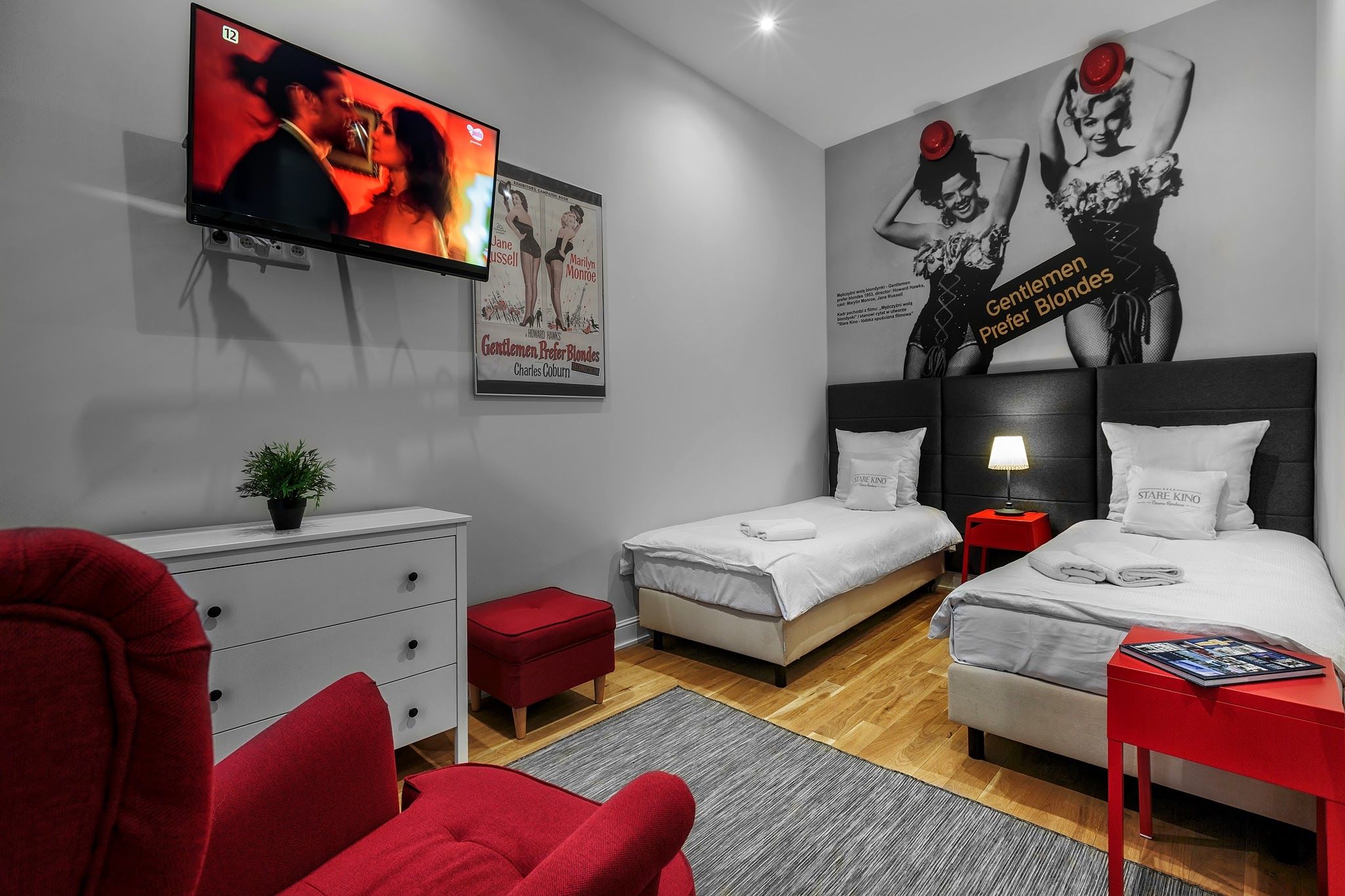A hotel room with two beds, a TV, a poster, a red chair, a white dresser, and a small red table.