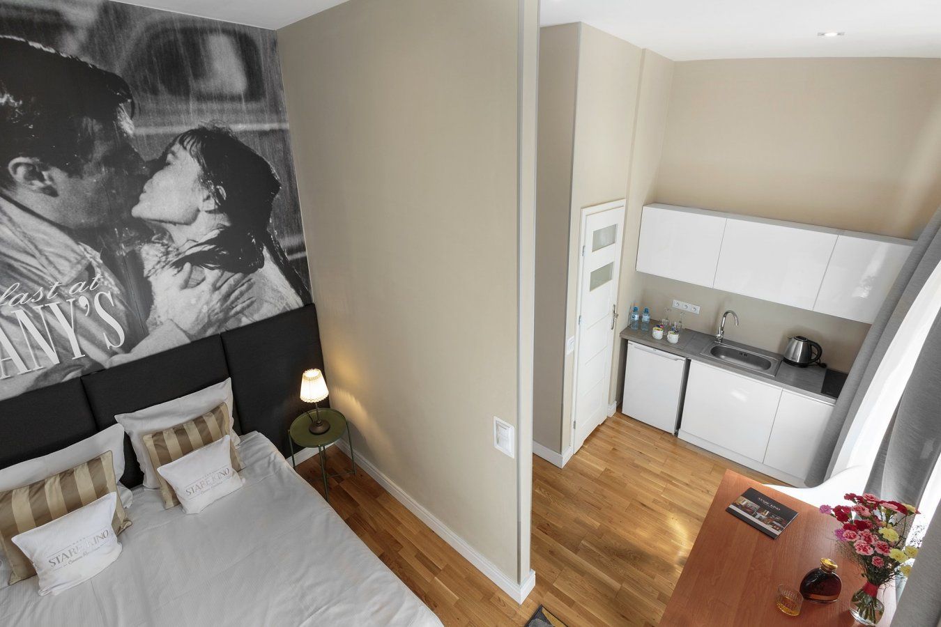 A bedroom with a bed, bedside table, lamp, and a wall with a poster. Adjacent is a kitchenette with a sink, stove, and cabinets.