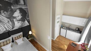 A bedroom with a bed, bedside table, lamp, and a wall with a poster. Adjacent is a kitchenette with a sink, stove, and cabinets.