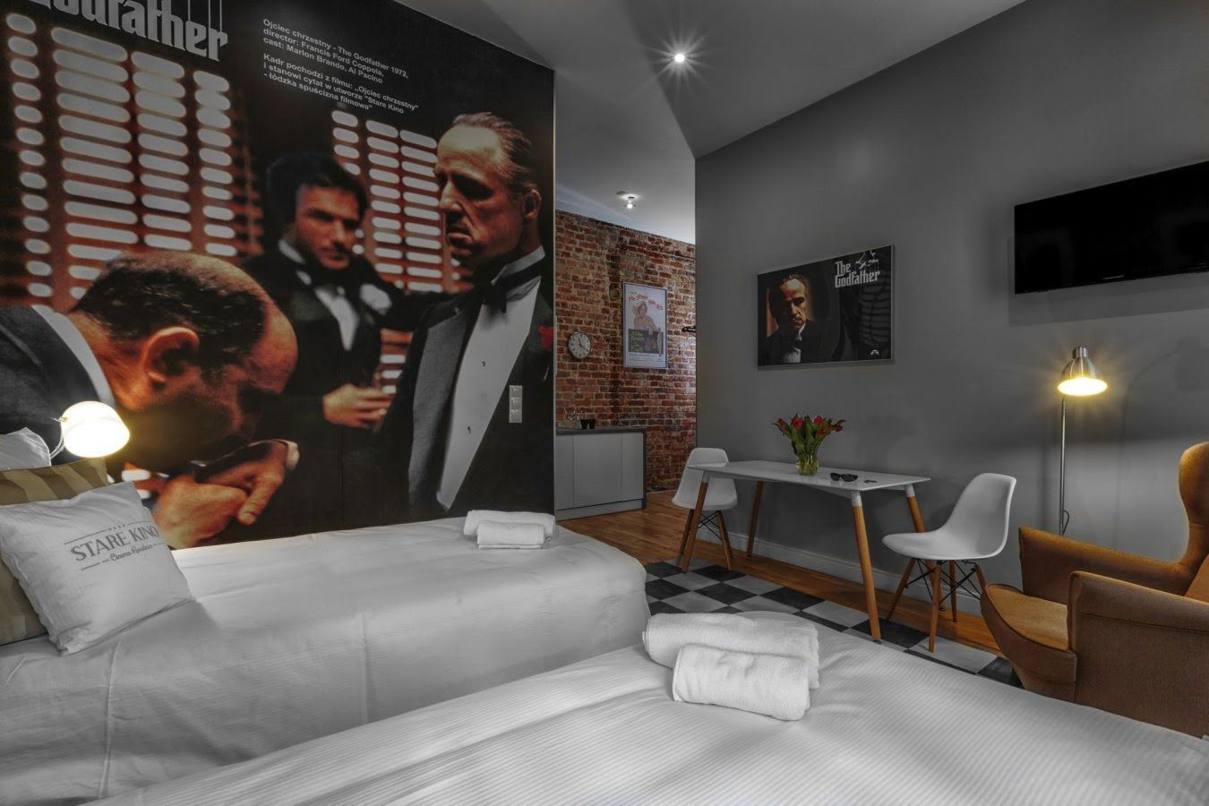 A modern hotel room with a large poster of 'The Godfather,' a flat-screen TV, a dining area with a table and chairs, and a sofa with towels.