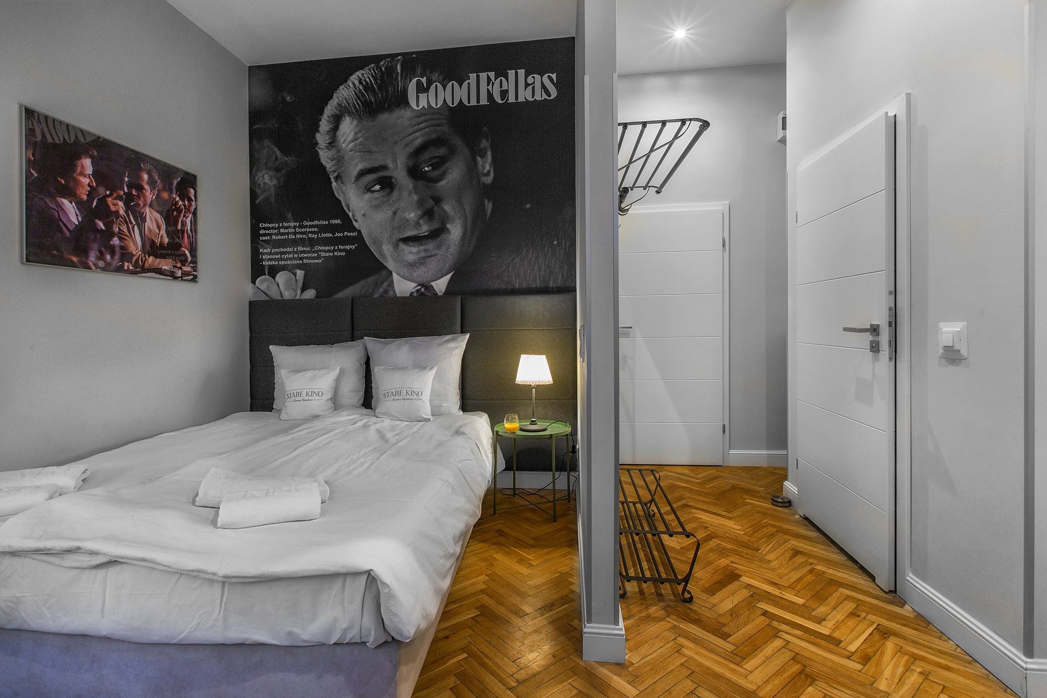 A hotel room with a bed, a nightstand, a lamp, a poster, and a wooden floor.