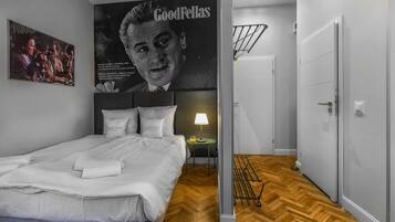 A hotel room with a bed, a nightstand, a lamp, a poster, and a wooden floor.