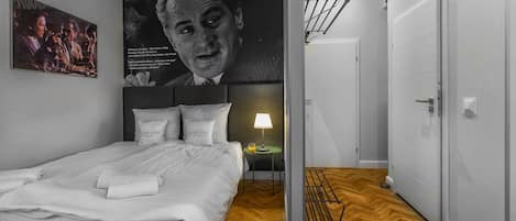 A hotel room with a bed, a nightstand, a lamp, a poster, and a wooden floor.