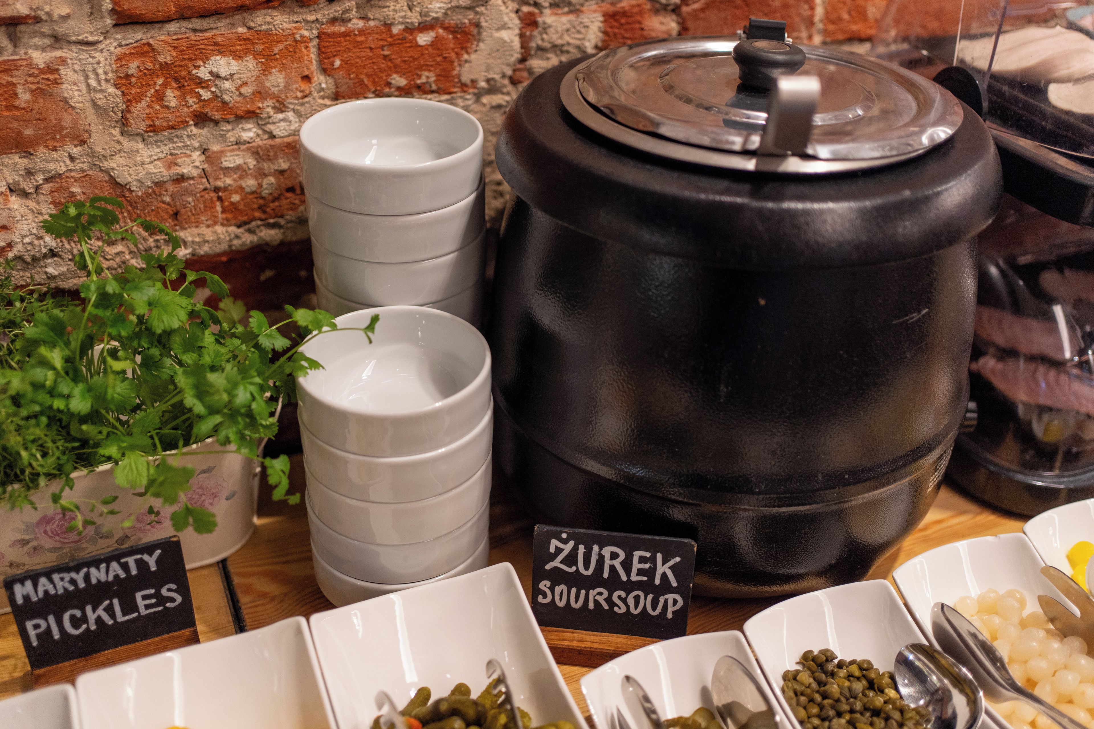 A buffet setup with labeled containers for pickles and sour soup, a large black pot, and a potted plant.