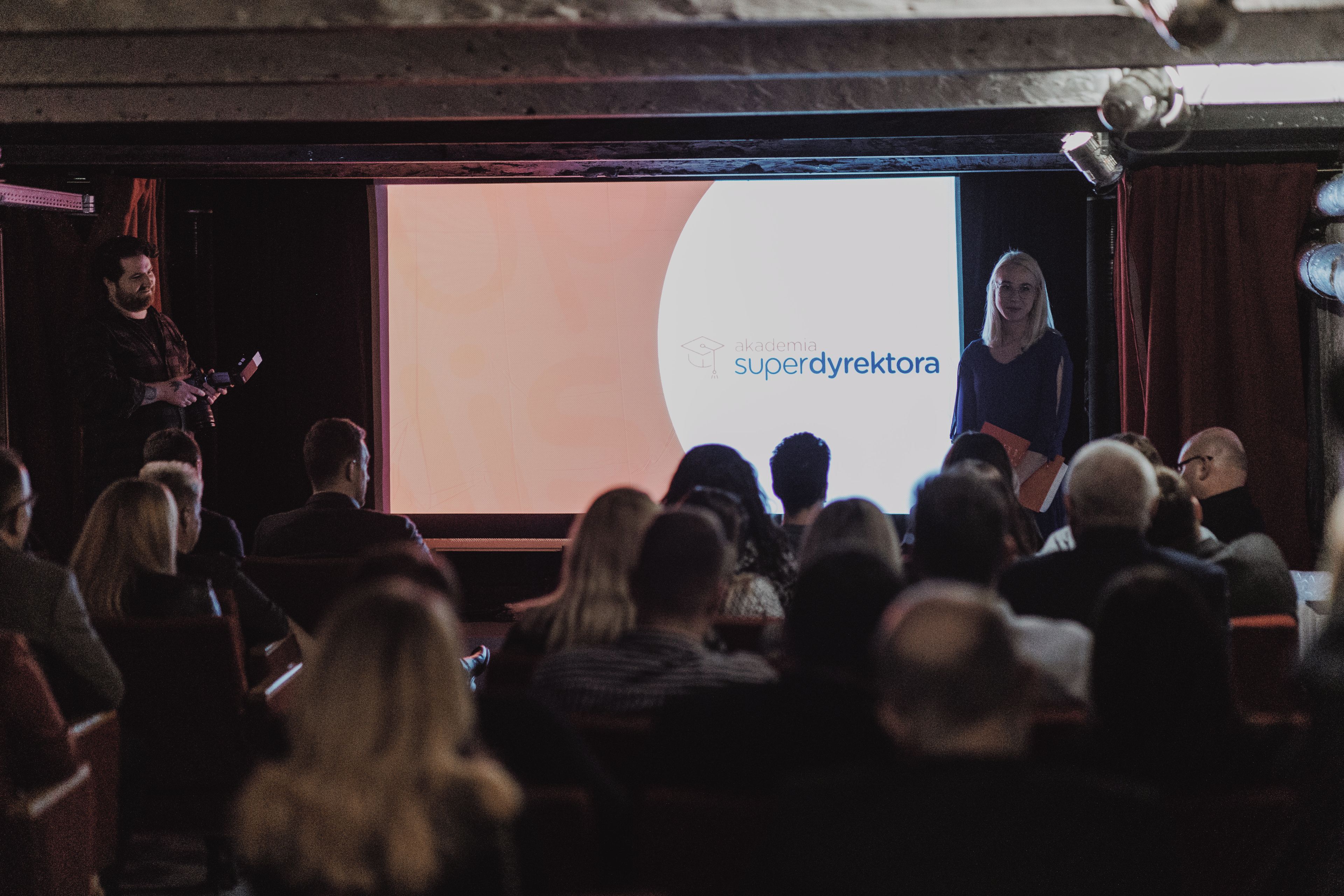 A presentation in a dimly lit room with a projector screen displaying 'akademia superdyerektora' and a seated audience.