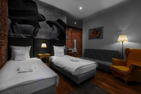 A hotel room with two beds, a sofa, a chair, a lamp, a brick wall, and a poster.