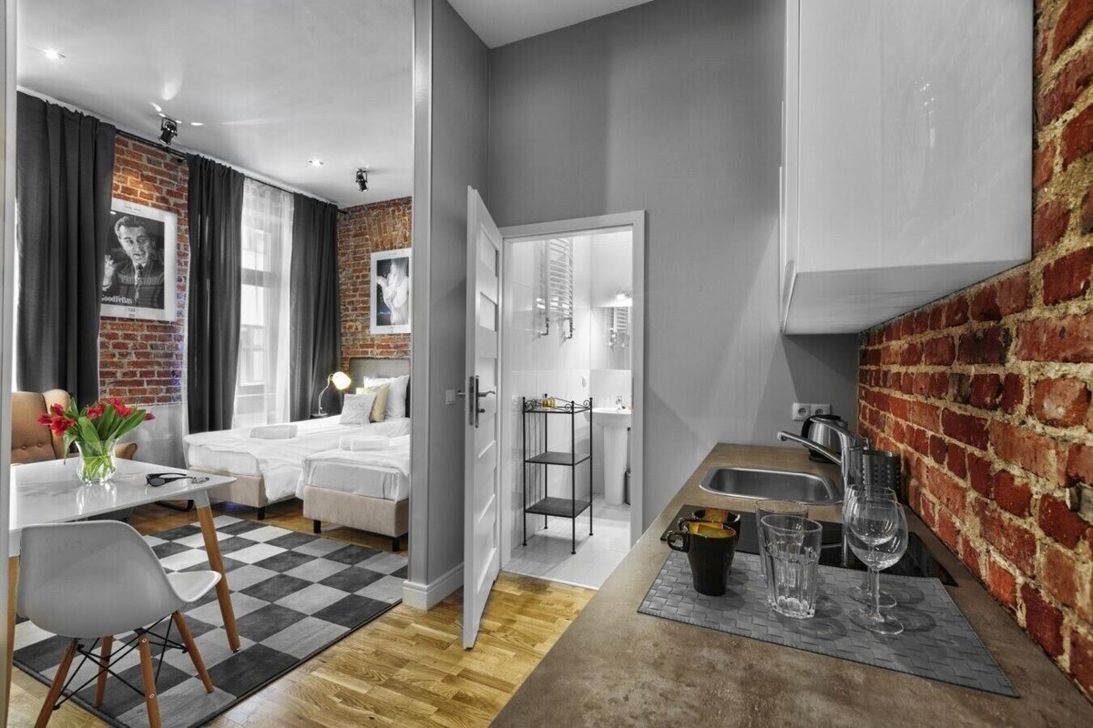 Ein modernes Hotelzimmer mit Schachbrettmuster-Boden, einem Bett, einem Schreibtisch und einer Küchenzeile mit Backsteinwand.