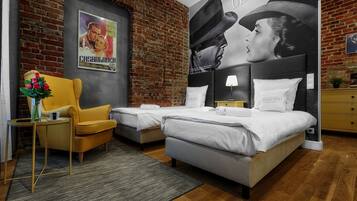 A hotel room with a brick wall, two beds, a yellow armchair, a small table with a vase of flowers, and a poster on the wall.