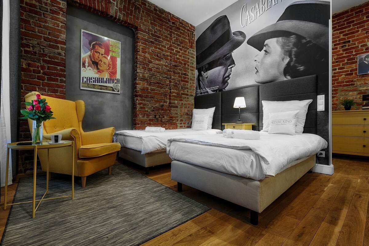 Ein Hotelzimmer mit Backsteinwand, zwei Betten, einem gelben Sessel, einem kleinen Tisch mit Blumenvase und einem Poster an der Wand.