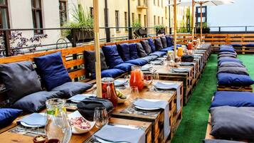 An outdoor dining area with long wooden tables, blue cushioned benches, and a green artificial turf floor.